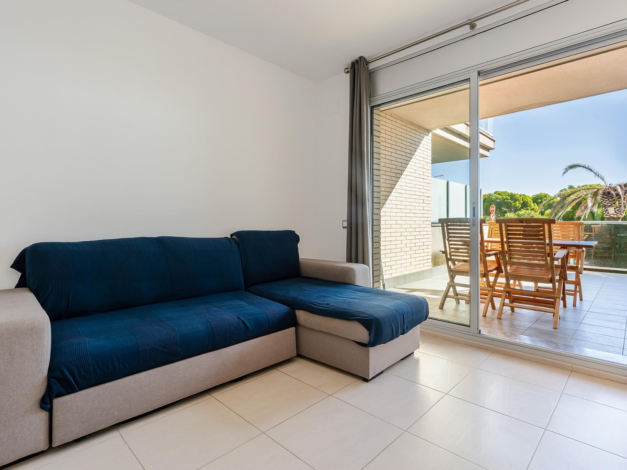 Een moderne woonkamer met een hoekbank en een glazen deur die leidt naar een terras met houten meubels.