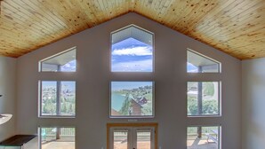 A spacious room with a high ceiling and multiple large windows offering views of a residential area and a body of water.