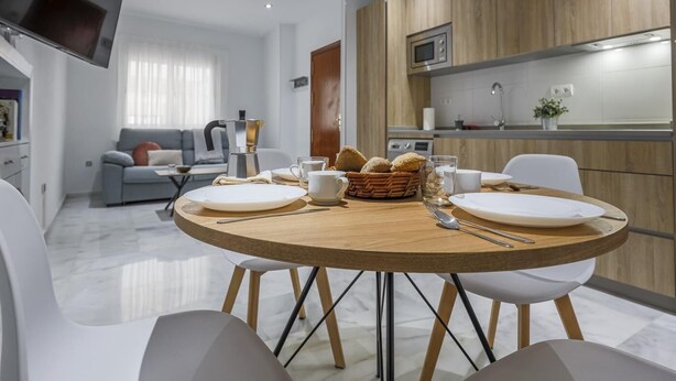 Una sala de estar moderna con una mesa de comedor redonda de madera, sillas blancas y un televisor de pantalla plana montado en la pared.