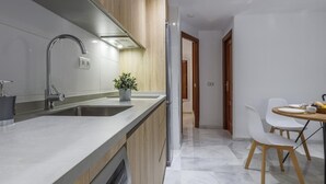 Una cocina moderna con lavavajillas integrado, un lavamanos de acero inoxidable y armarios de madera. Hay una zona de comedor con una mesa redonda y dos sillas.