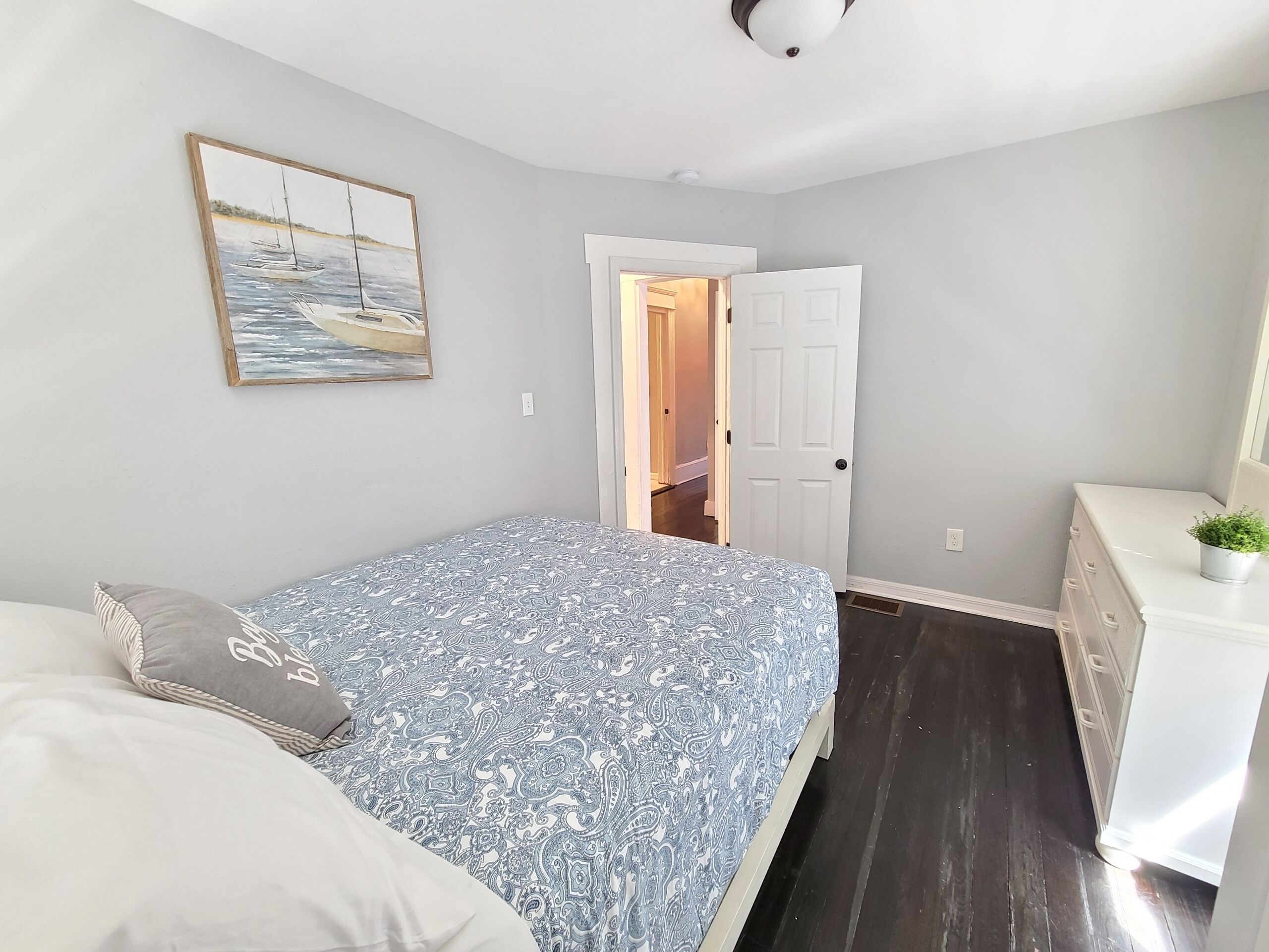 A bedroom with a bed, a dresser, a door leading to another room, and a painting on the wall.