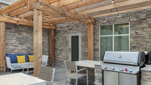 Un patio exterior con paredes de piedra, una pérgola de madera, una parrilla y un área de estar con mesa y sillas.