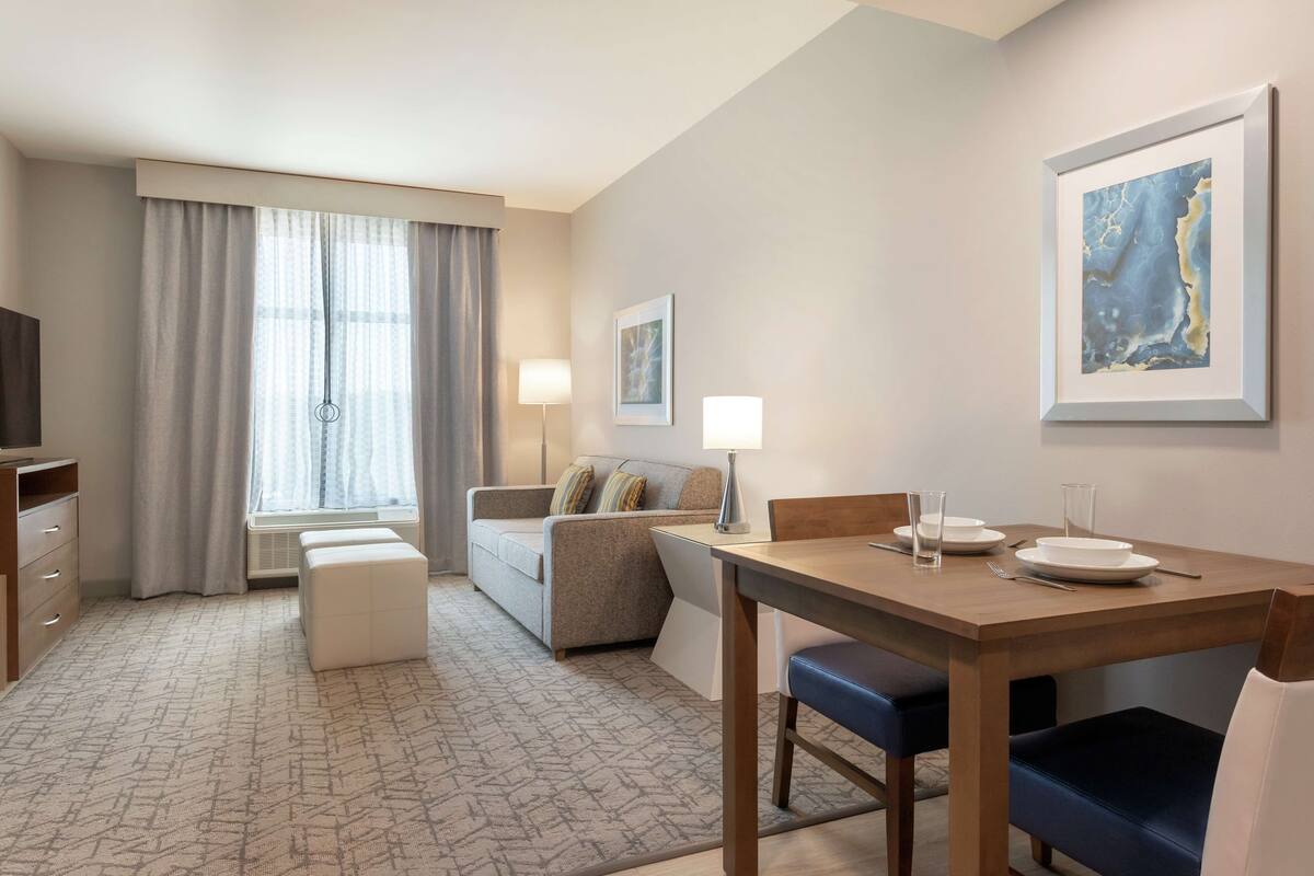 Habitación de hotel con zona de comedor, sofá, televisión y un gran ventanal con cortinas.