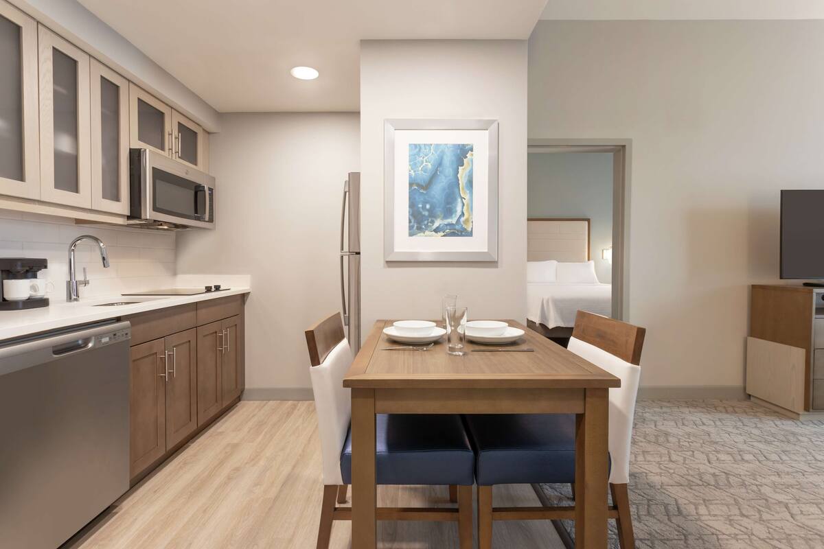 Una cocina moderna con armarios de madera, un refrigerador de acero inoxidable y un microondas. Hay una mesa de comedor con sillas y una obra de arte enmarcada en la pared.