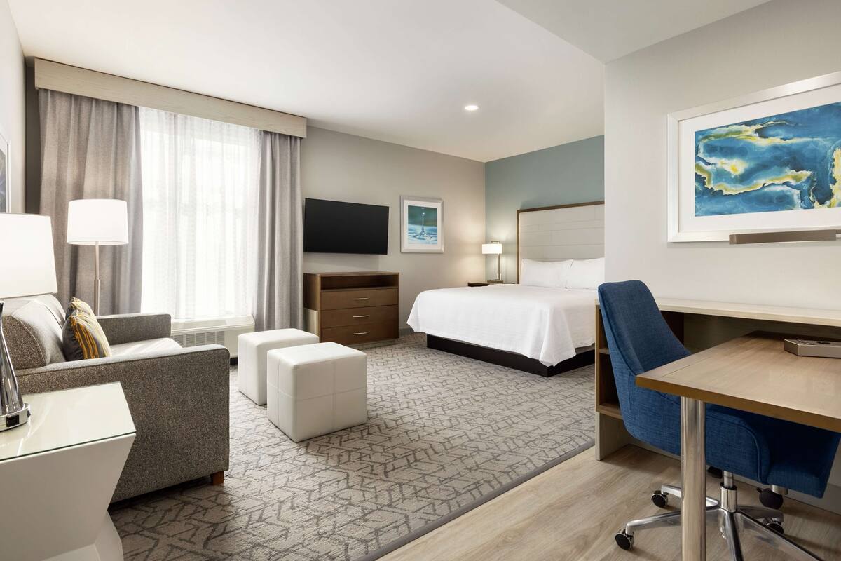 Una habitación de hotel con cama, escritorio, silla, sofá, televisor y obras de arte en las paredes.