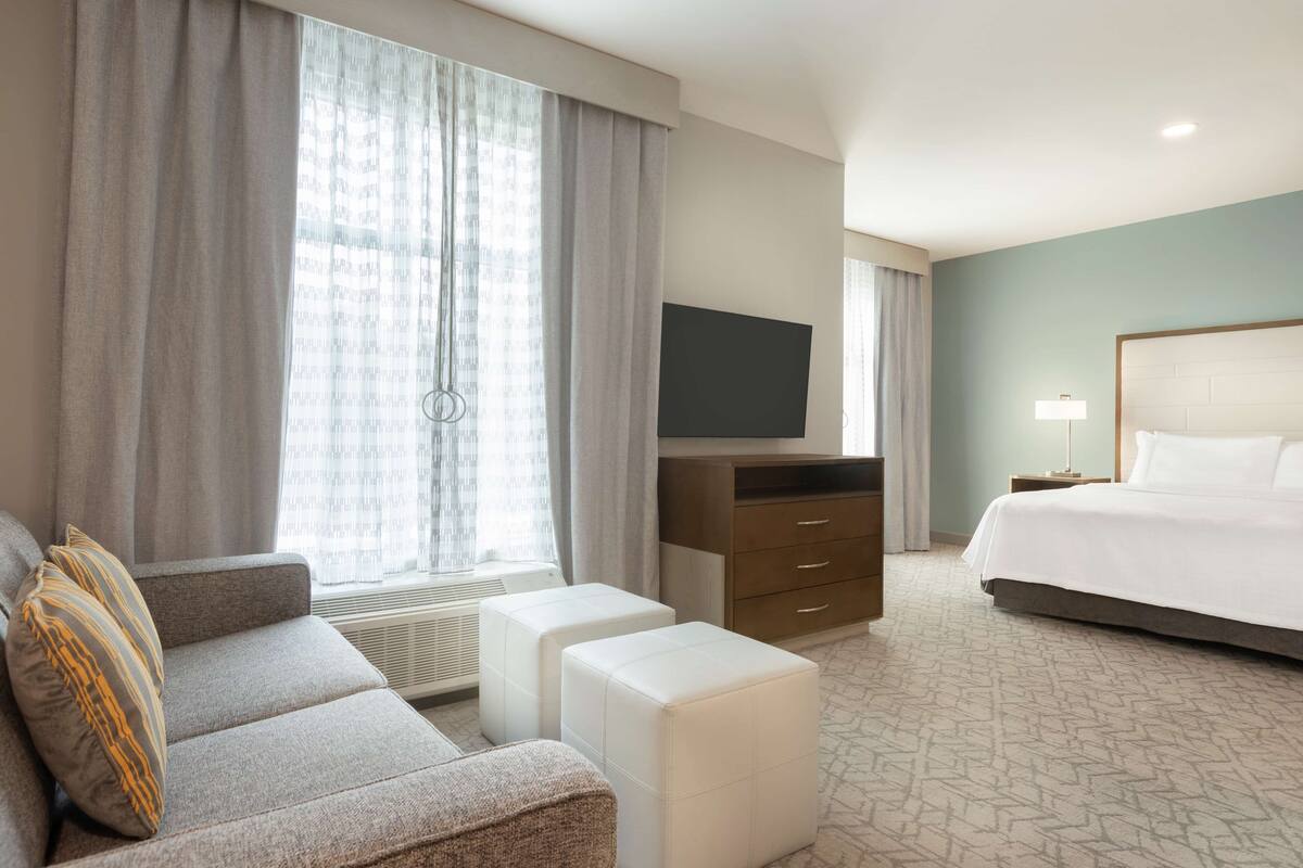 Habitación de hotel con sofá, otoman, televisor de pantalla plana, cama y mesita de noche con lámpara.