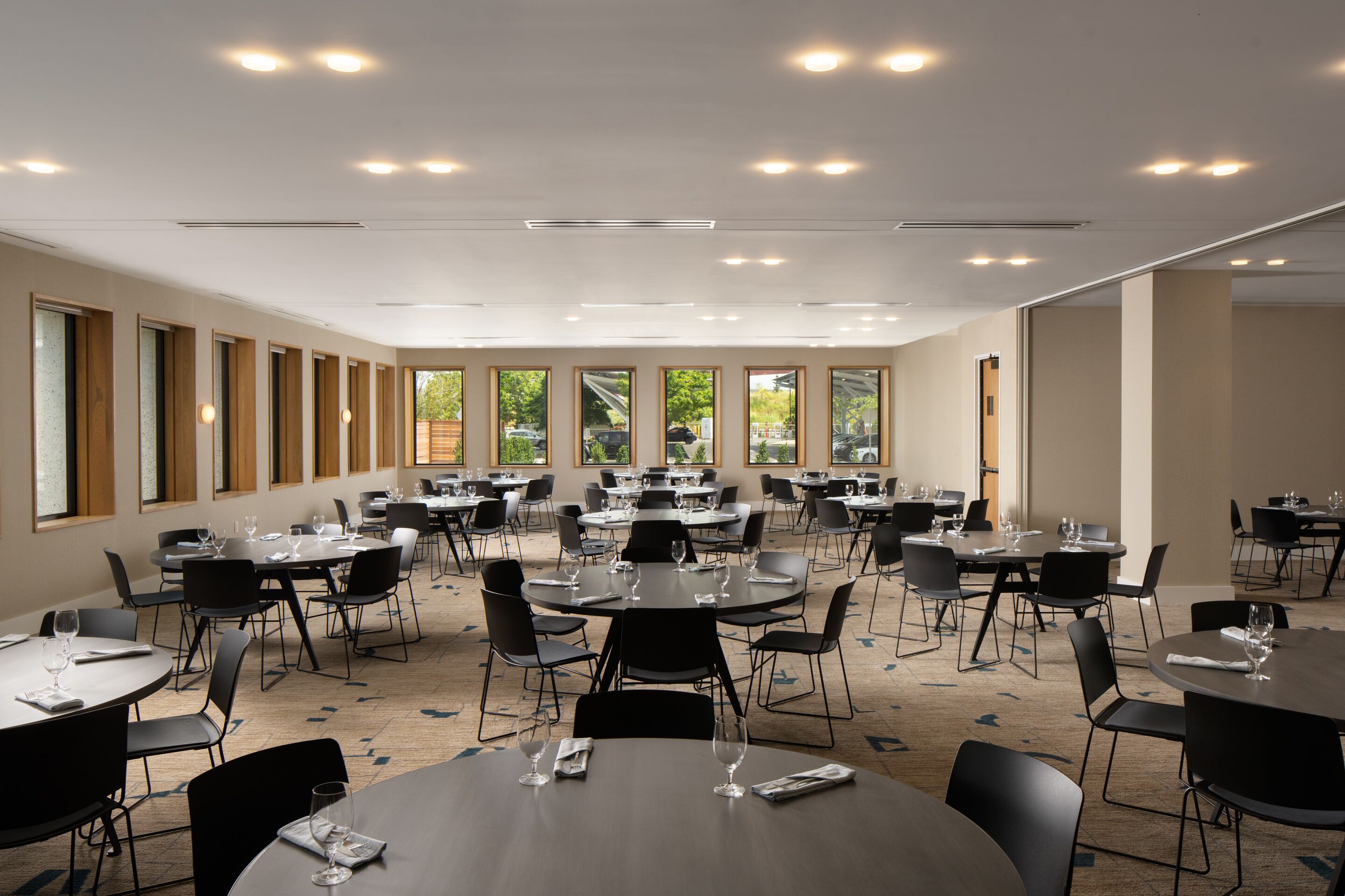 Een ruime congreszaal met meerdere ronde tafels en stoelen, grote ramen met uitzicht op groen en inbouwspots.