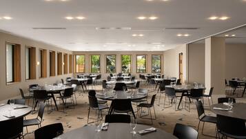 Een ruime congreszaal met meerdere ronde tafels en stoelen, grote ramen met uitzicht op groen en inbouwspots.