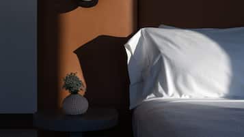 A neatly made bed with white linens, a small round table with a white vase and a plant, and a wall-mounted light fixture.