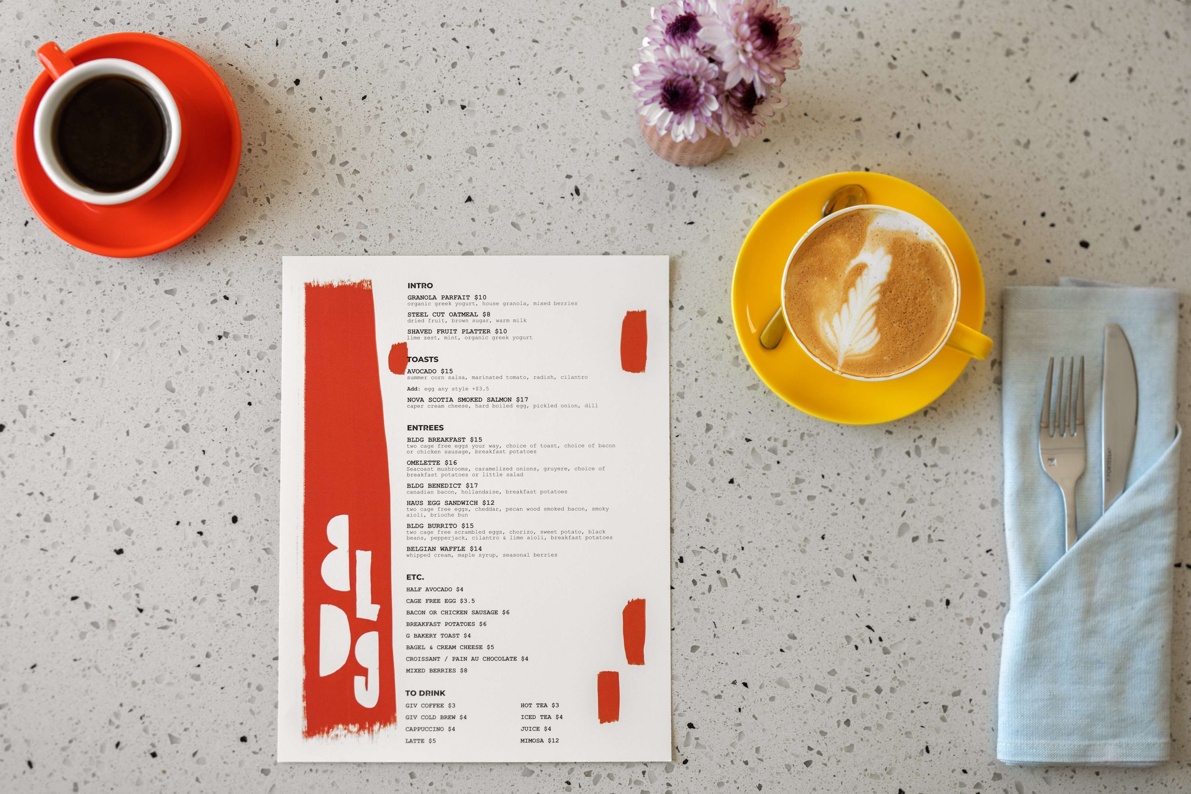 A menu with various food and drink options, a cup of coffee, a small vase with flowers, and a napkin with cutlery on a speckled countertop.