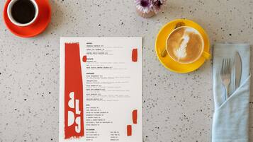 A menu with various food and drink options, a cup of coffee, a small vase with flowers, and a napkin with cutlery on a speckled countertop.