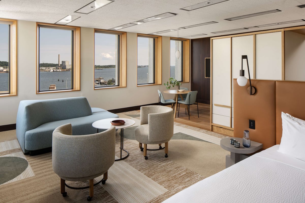 A hotel room with a bed, a sofa, two armchairs, a small table, and a view of the water and city skyline.