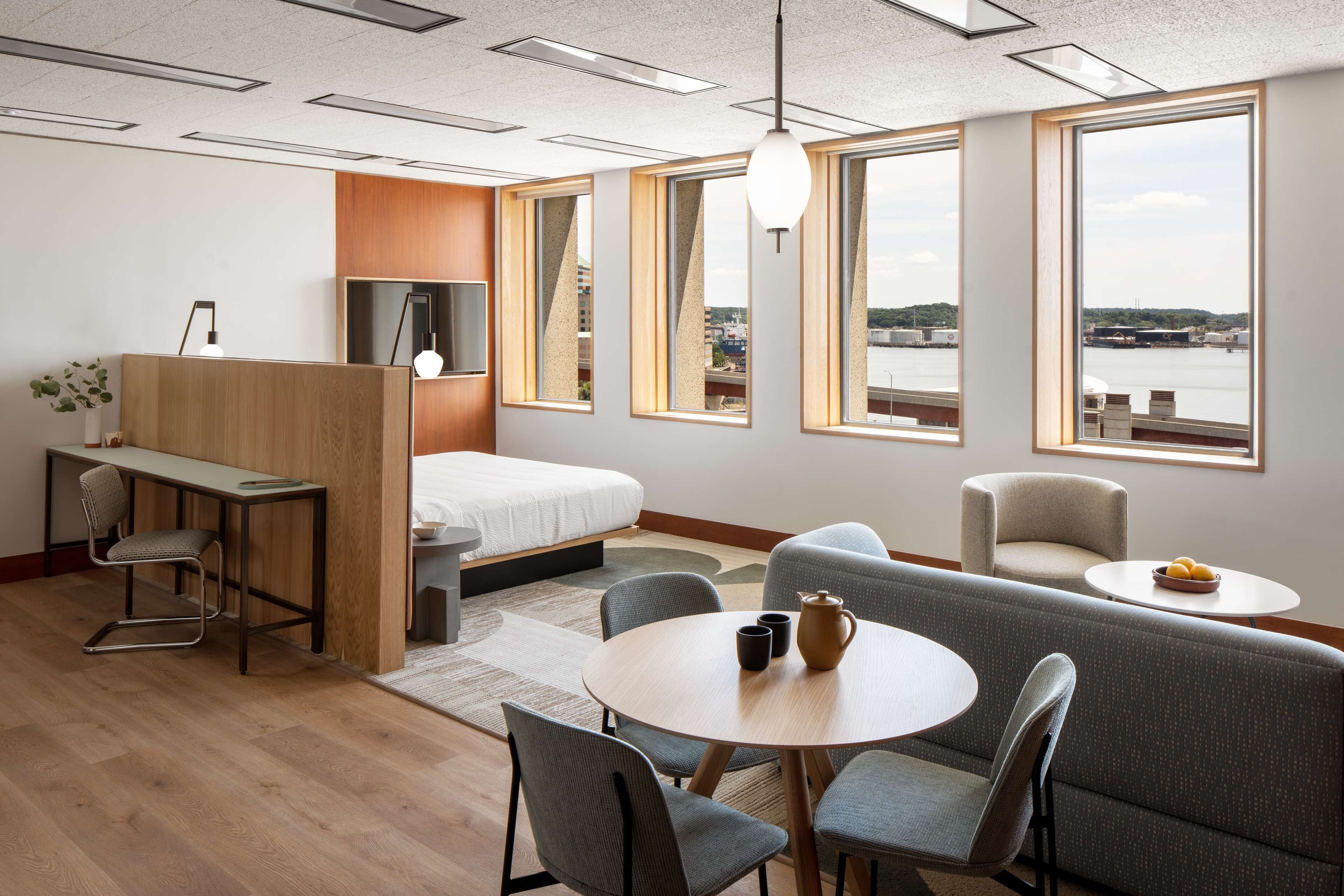 Ein modernes Hotelzimmer mit einem Holztisch, einem Bett, einer Couch, einem Esstisch und großen Fenstern mit Blick auf das Wasser.