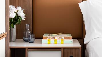 A neatly arranged bedside table with a vase of white flowers, two clear glasses, and a stack of books titled 'JENS RISOM'.
