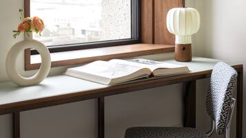 A desk with a book, a vase with flowers, and a lamp by a window.