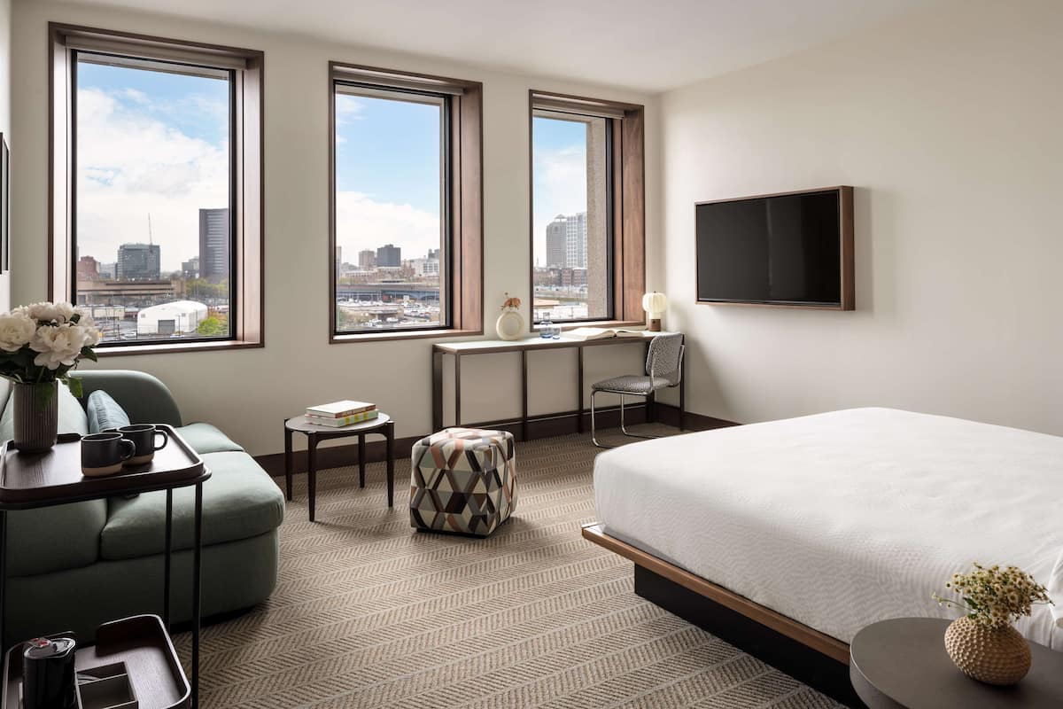 A hotel room with a large bed, a sofa, a small table, a chair, and a city view through large windows.