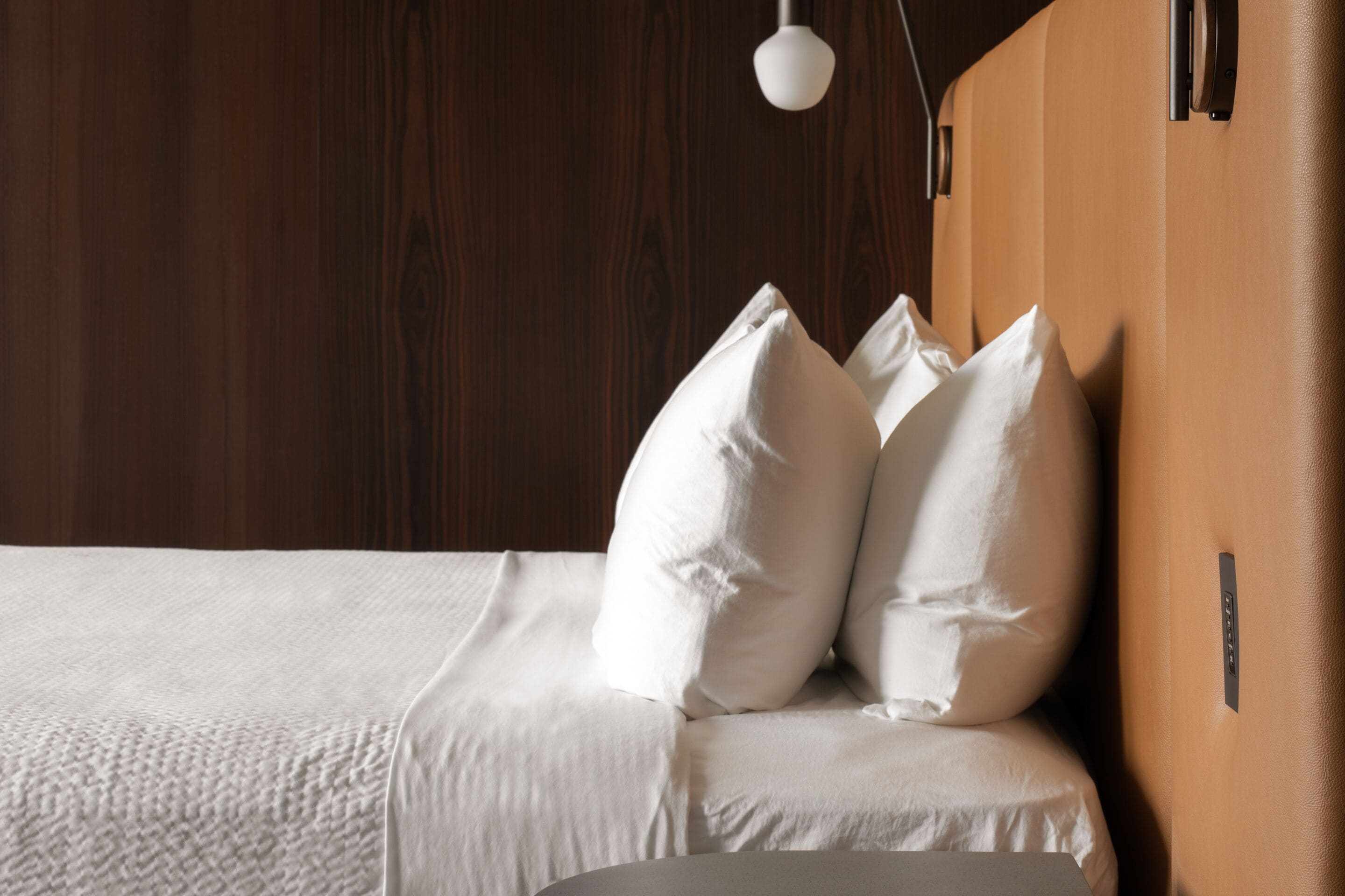 Cama bem arrumada com lençóis e almofadas brancas, cabeceira de madeira e luminária pendente.