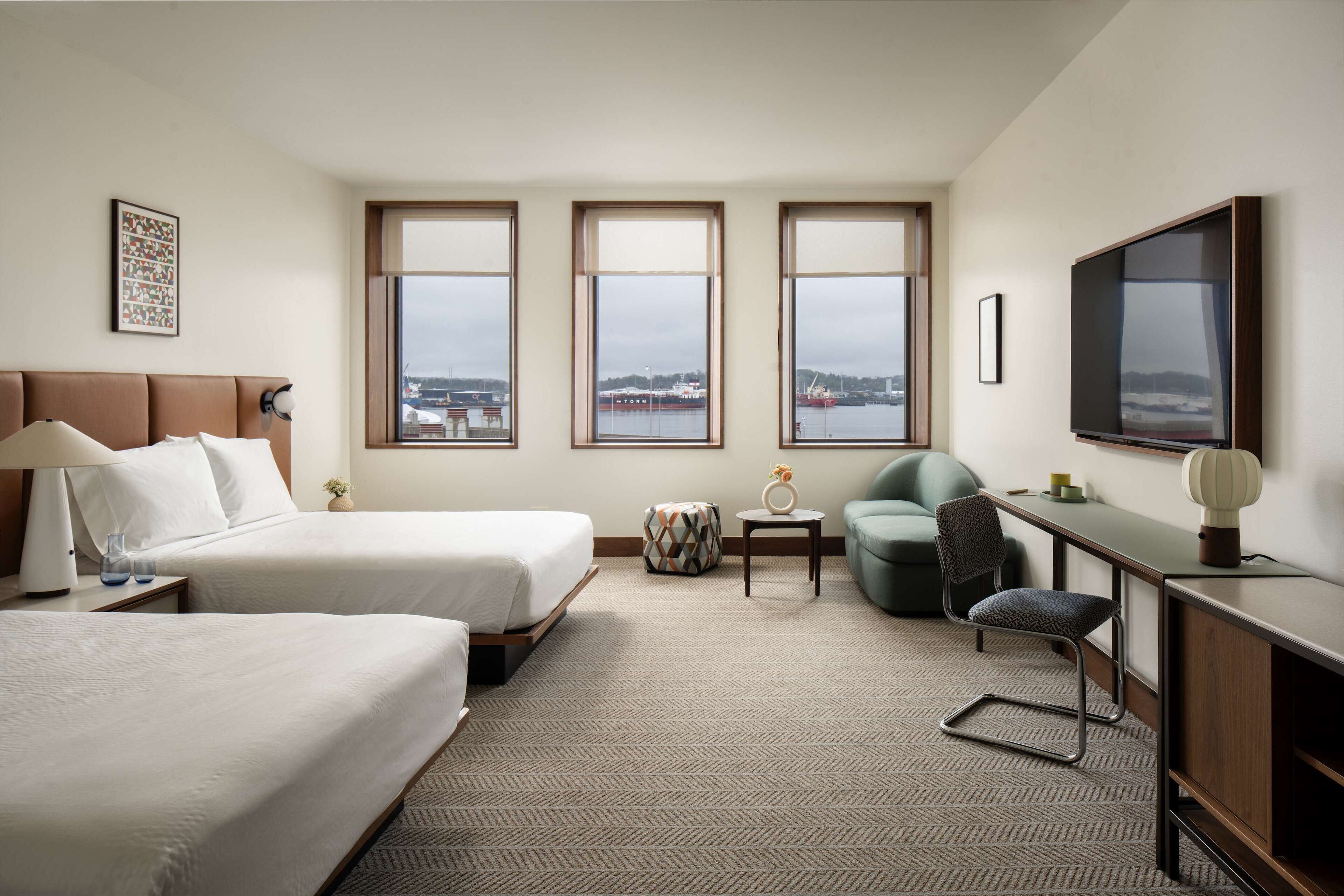 A hotel room with two beds, a desk, a chair, a TV, and large windows with a view of the city.