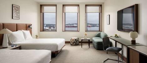 A hotel room with two beds, a desk, a chair, a TV, and large windows with a view of the city.
