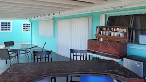 A room with a wooden table and chairs, a radiator, a window with curtains, and a shelf with bottles. - Maryville Studio (St.Thomas)