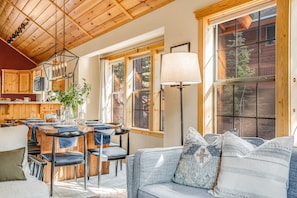 A cozy interior with a dining area, wooden ceiling, large windows, and a sofa with patterned cushions.