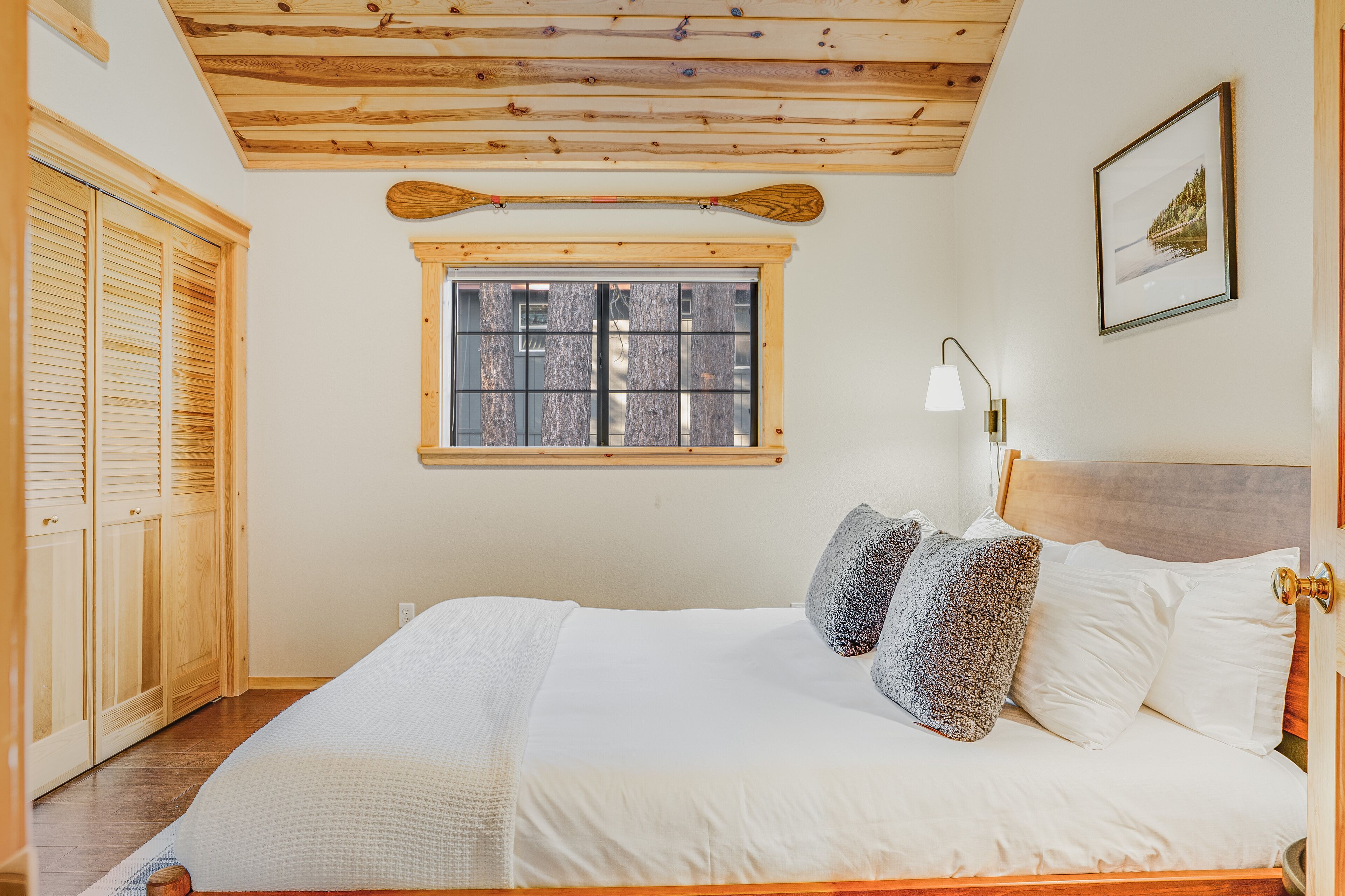 A bedroom with a wooden ceiling, a window with a view, a bed with white bedding, and a wooden wardrobe.
