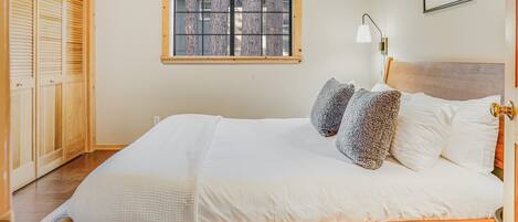 A bedroom with a wooden ceiling, a window with a view, a bed with white bedding, and a wooden wardrobe.