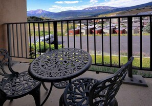 A balcony with a metal table and chairs, overlooking a parking area and buildings.