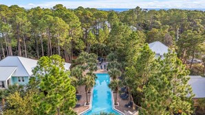 A resort with a central swimming pool surrounded by tall pine trees, multiple buildings with sloped roofs, and a clear view of the ocean in the background. - Bring Fido! Stylish 4BR Home in Eastern Lake (Santa Rosa Beach)