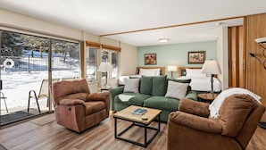 A cozy living room with a sofa, armchairs, and a coffee table. There is a bed in the background and a view of the outdoors through large windows. - Steps To Snowmass Base Village! Hot Tub, Pool & Mountain Access! Patio W/Grill, Covered Parking (Snowmass Village)