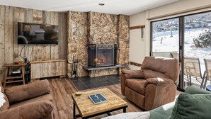 A cozy living room with a stone fireplace, leather sofas, a flat-screen TV, and a view of a snowy landscape. - Steps To Snowmass Base Village! Hot Tub, Pool & Mountain Access! Patio W/Grill, Covered Parking (Snowmass Village)