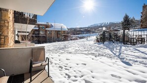 A snow-covered outdoor area with a chair and a table, a stone building, and a view of a mountain range. - Steps To Snowmass Base Village! Hot Tub, Pool & Mountain Access! Patio W/Grill, Covered Parking (Snowmass Village)