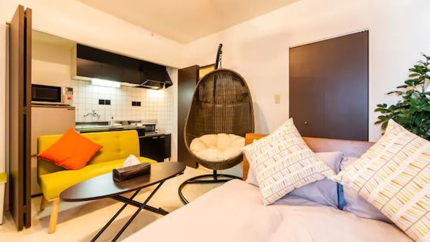 A compact living space with a kitchenette, a sofa with orange cushions, a hanging chair, and a bed with patterned pillows. - ORANGE (Sapporo)