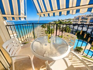 Un balcon avec une table en verre, des chaises blanches et une vue sur la mer et les bâtiments.