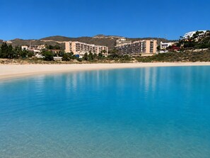 Una piscina azul cristalina con una playa arenosa y edificios al fondo.