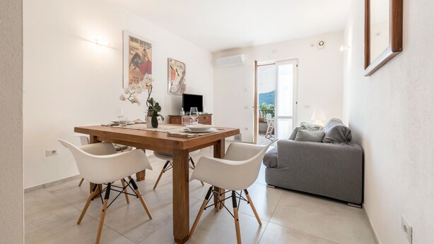 A dining area with a wooden table, white chairs, and a grey sofa in a bright room with white walls and a view through an open door. - Appartamento "Le Cementine" with Lake View, Mountain View, Wi-Fi, A/C & Terrace (Vesio, Tremosine sul garda)