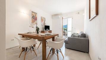 Uma sala de jantar com mesa de madeira, cadeiras brancas e sofá cinza, num espaço iluminado com paredes brancas e vista através de uma porta aberta.