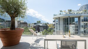 A terrace with a large potted olive tree, two white chairs, a table with a bottle and glasses, and a view of buildings and mountains in the background. - Aris Apartments & Sky Pool - Tonelli Hotels - Adults Only (Riva del Garda)
