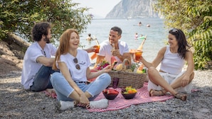 A group of people sitting on a beach with a picnic setup, including a basket and bowls of fruit. - Aris Apartments & Sky Pool - Tonelli Hotels - Adults Only (Riva del Garda)