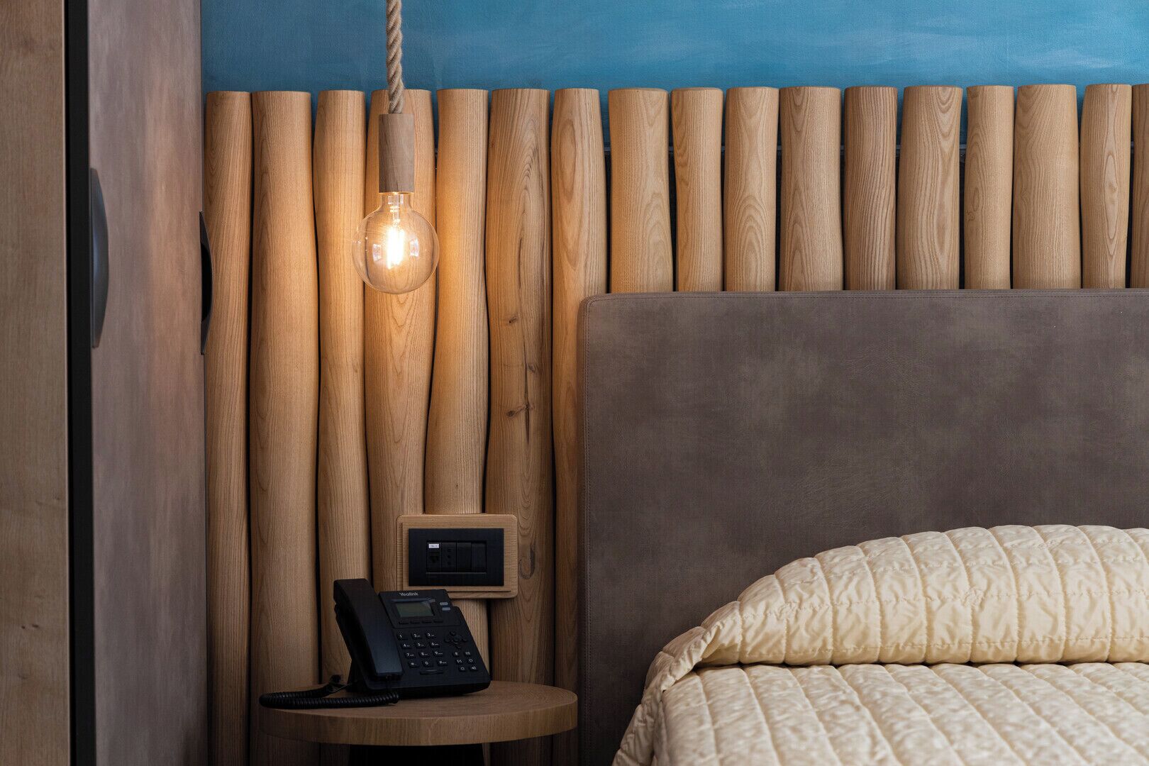 A bedroom with a wooden slat wall, a round bedside table with a telephone, and a bed with a quilted cover.