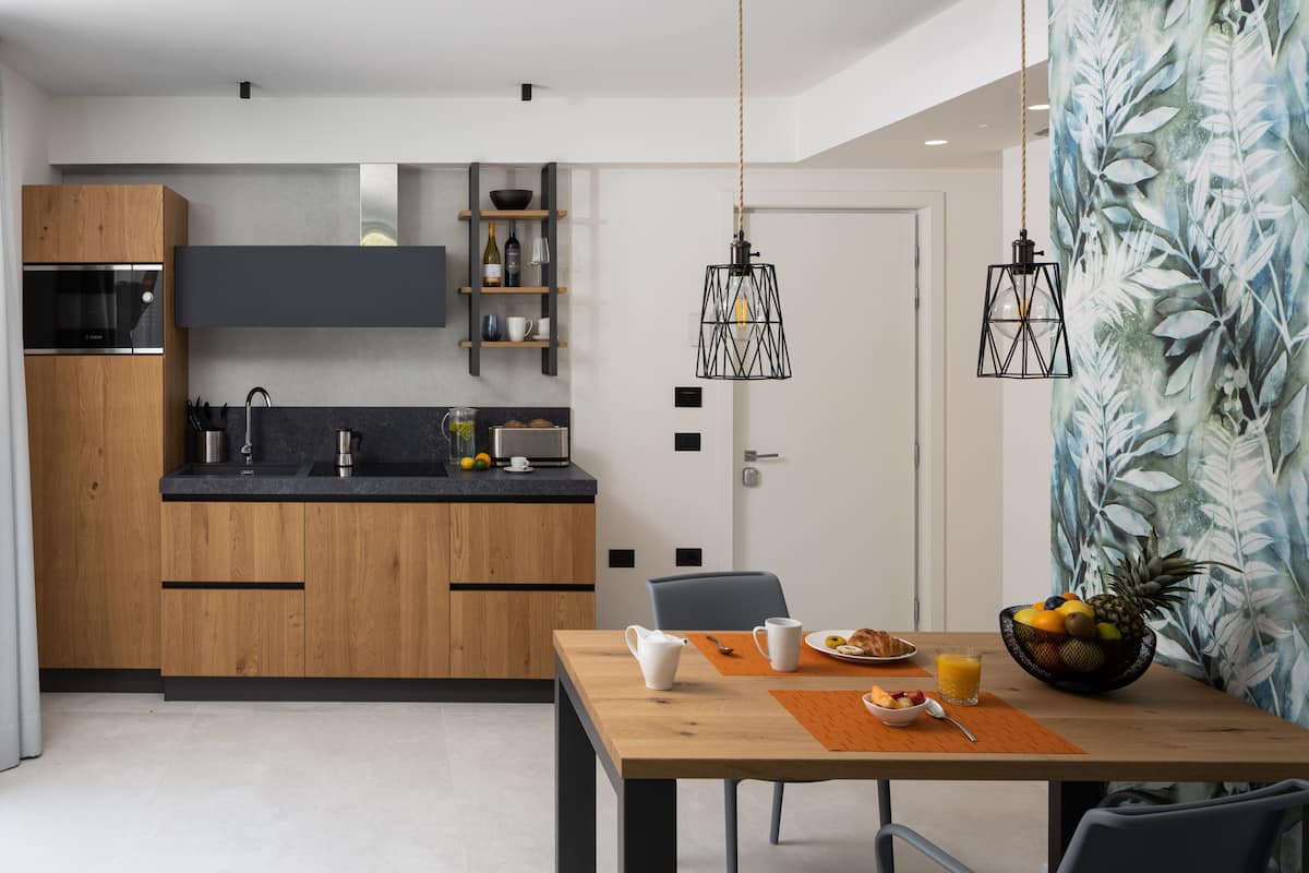 Een moderne keuken met houten kastjes, een ingebouwde oven en een zwart aanrecht. Er is een eethoek met een houten tafel, stoelen en een schaal fruit.