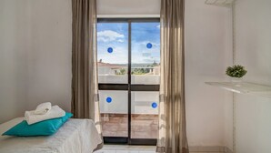 A room with a bed, a window with curtains, a balcony with a view, and a small plant on a shelf. - Vila Casa do Cerro with Pool, Garden & Wi-Fi (Boliqueime)