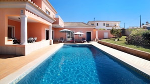 Uma piscina com borda azulejada, um alpendre coberto com espreguiçadeiras e um edifício com telhado em azulejos.