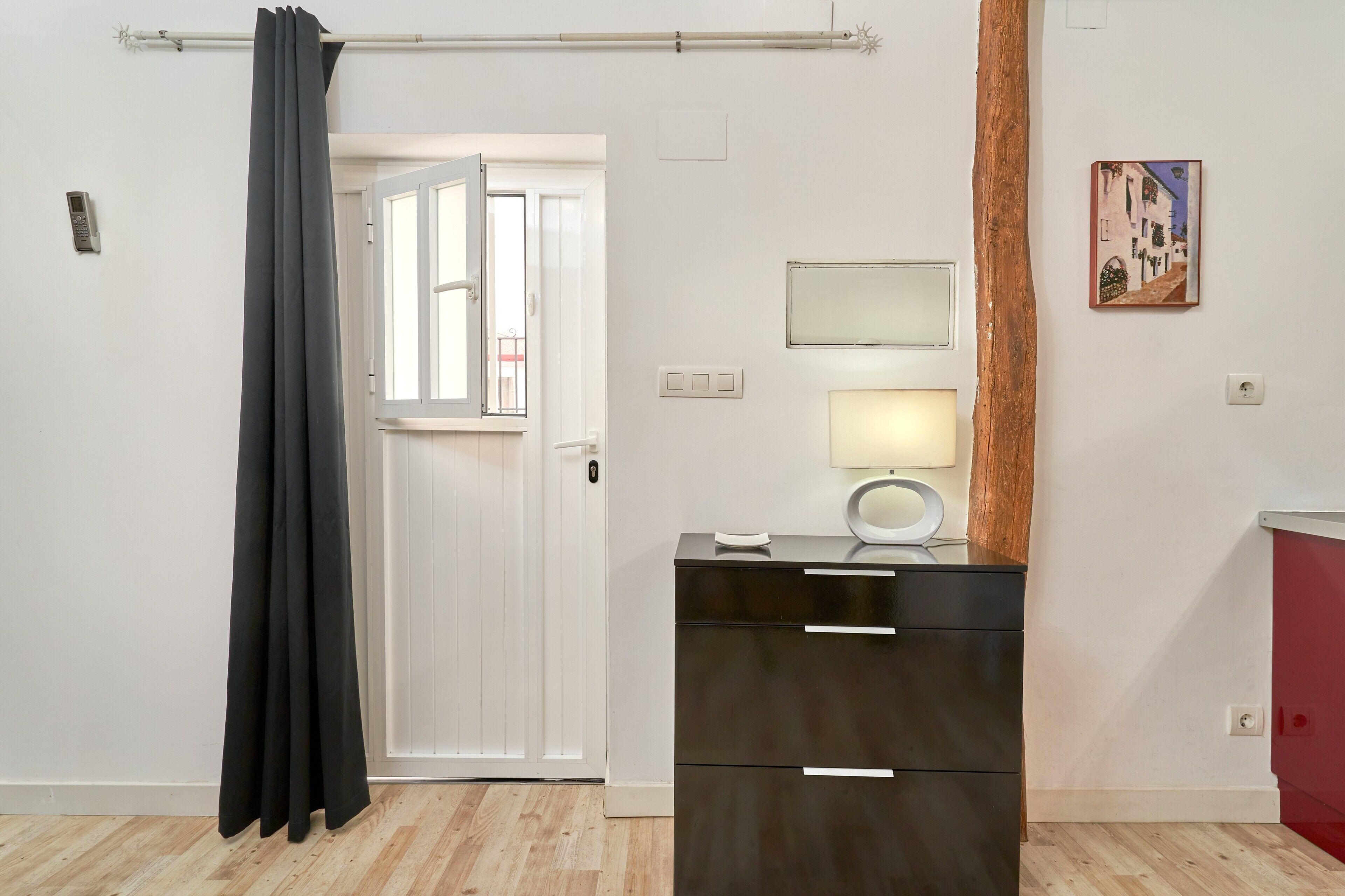A room with a white door, a black cabinet with drawers, a lamp, and a painting on the wall.