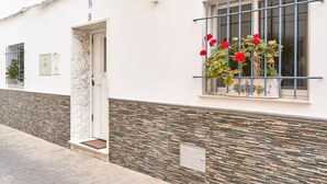 Un bâtiment à la façade blanche, avec une porte, une fenêtre ornée d’une grille métallique et un jardinier planté de fleurs rouges.