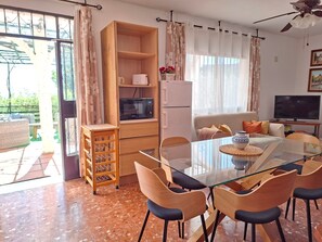Ein modernes Wohnzimmer mit einem Glastisch, Holzstühlen, einer Couch, einem Fernseher und einer Küchenzeile inklusive Mikrowelle und Kühlschrank.