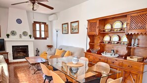 A dining area with a glass table, wooden chairs, a fireplace, and a kitchen with wooden cabinets and decorative plates. - Holiday Home 'Sailor House Slow Life In Nature' with Mountain View, Wi-Fi and Air Conditioning (Málaga)