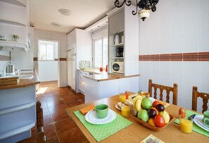 Een moderne keuken met witte kasten, een eettafel gedekt met fruit en een groen kopje, en een raam met jaloezieën.