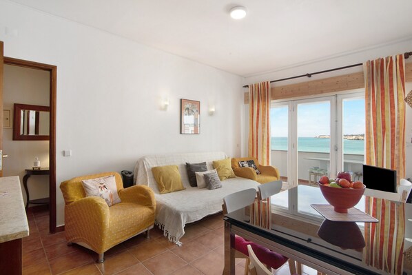 A living area with a sofa, a dining table with chairs, a TV, and a view of the sea. - Apartment 'Caravela' with Sea View, Balcony & Wi-Fi (Armação de Pêra)