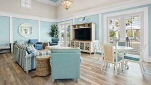 Un salon spacieux comprenant un canapé d’angle, un téléviseur et un coin repas avec des chaises et une table blanches.